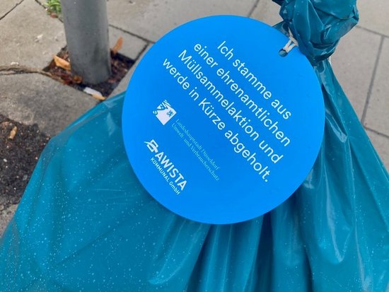 Ein blauer Müllbeutel am Straßenrand trägt einen runden blauen Anhänger. Dieser besagt: „Ich stamme aus einer ehrenamtlichen Müllsammelaktion und werde in Kürze abgeholt.“ Darunter befinden sich die Logos des Landschaftsverbandes Düsseldorf und der AWISTA.