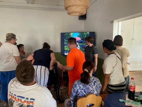 Eine Gruppe von Menschen steht in einem hellen Raum und schaut gemeinsam auf einen Fernseher, auf dem ein Videospiel läuft. Eine Person im Rollstuhl ist Teil der Gruppe.