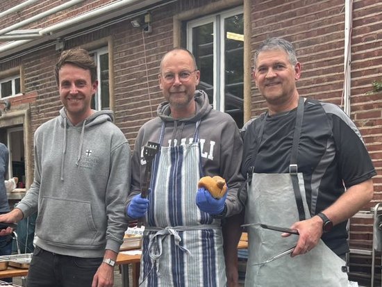 Drei Männer stehen lächelnd hinter einem langen Grill, auf dem Würstchen und Burger zubereitet werden. Der Mann in der Mitte hält ein Brötchen, der Mann rechts Grillzangen. Im Hintergrund sind ein Backsteingebäude und weitere Personen zu sehen.
