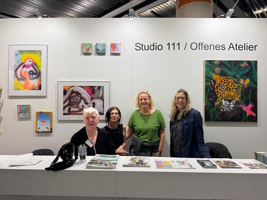 Vier Frauen stehen hinter einem weißen Tisch an einem Messestand mit der Aufschrift „Studio 111 / Offenes Atelier“. An der Wand hängen diverse farbenfrohe Gemälde und Kunstwerke. Auf dem Tisch liegen Informationsbroschüren.