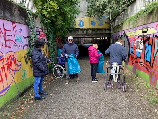 Vier Menschen, darunter eine Person mit Rollator und ein Mann mit blauem Müllsack, sammeln Müll in einem mit buntem Graffiti besprühten Unterführung. Die Wände sind teilweise mit Efeu bewachsen.