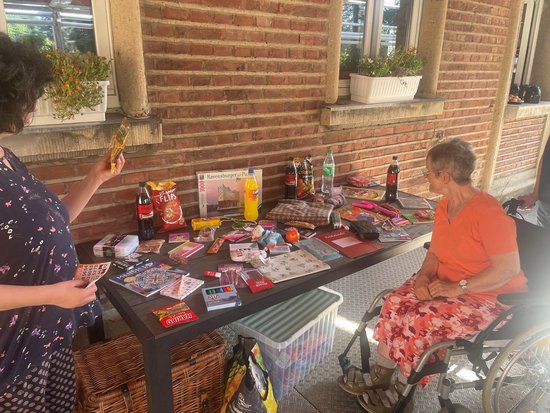 Eine Frau steht vor einem Tisch, der mit Süßigkeiten, Getränken und einem Puzzle beladen ist. Eine ältere Dame im Rollstuhl sitzt ihr gegenüber. Auf dem Tisch liegen diverse Artikel, darunter Cola, Chips und Buntstifte.