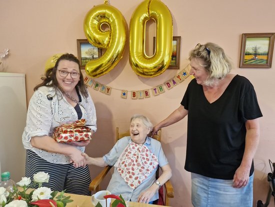 Eine ältere Frau feiert ihren 90. Geburtstag. Sie sitzt lachend zwischen zwei Frauen. Eine reicht ihr ein Geschenk, die andere steht lächelnd daneben. Goldene Ballons mit der Zahl 90 und eine Girlande schmücken den Hintergrund.
